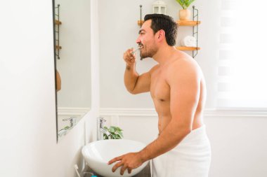 Side view of a shirtless young man using a toothbrush and brushing his teeth looking at the mirror after a shower in the bathroom