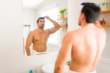 Hispanic muscular man using a comb and doing a stylish hairstyle in his hair while grooming after a shower in the bathroom