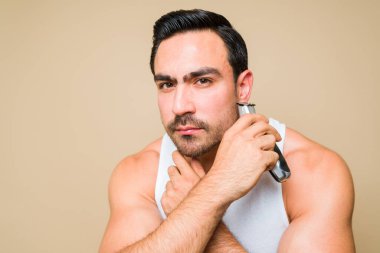 Attractive caucasian man trimming his beard and using an electric shaver while grooming in front of a studio yellow background
