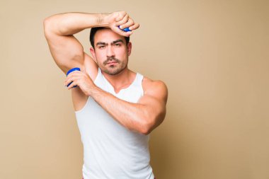 Athletic young man looking at the camera while using deodorant on his armpit to prevent sweating against a studio background