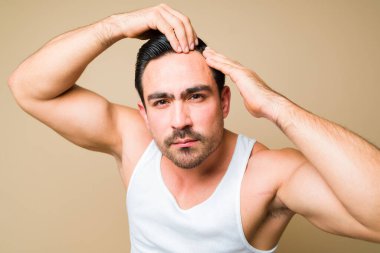 Stressed young man looking worried and concerned about baldness and hair receding while touching his hair against a studio background