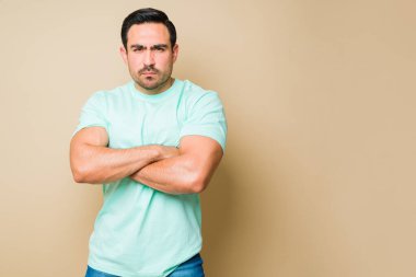 Angry man in his 20s looking upset and annoyed while arguing with a person and crossing his arms against a background with copy space