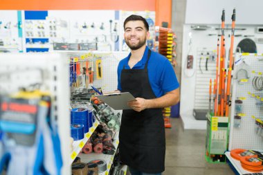 Cheerful retail worker with a blue uniform smiling holding a clipboard for inventory and looking happy to work at the hardware store 