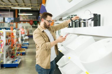 Handsome man texting his partner on the phone while shopping for a new bathroom faucet at the hardware store