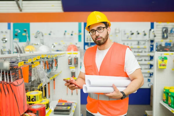 Handsome male engineer with a security helmet and vest holding blueprints while shopping at the hardware store