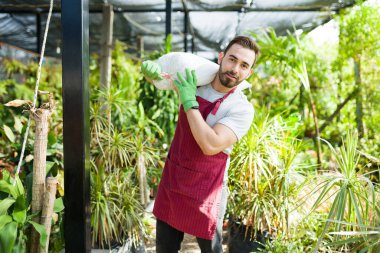 Strong caucasian worker carrying a bag of fertilizer soil to grow beautiful plants and flowers at the nursery garden