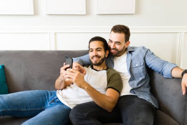 Cheerful gay couple hugging and using the smartphone to send text messages and look at social media while relaxing on the sofa at home 