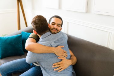 Attractive gay man in love embracing his partner and sharing their feelings while resting together at home in the living room