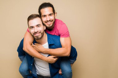 Fun caucasian gay man piggybacking his latin boyfriend looking happy against a studio background with copy space