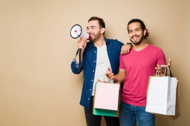 Smiling hispanic man looking happy while showing his shopping bags while his partner uses a megaphone to shout about a sale