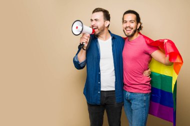 Cheerful attractive gay couple using a rainbow flag shouting with a megaphone during a protest to support LGBT rights 
