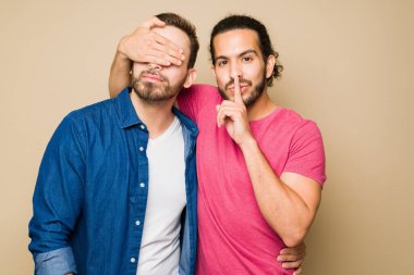 Latin gay young man covering his partner's eyes and asking to keep a secret about his lies or cheating affairs