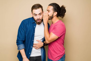 Handsome latin man whispering a secret in his boyfriend's ear looking surprised about the shocking news