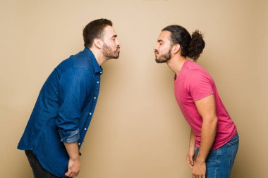 Side view of a caucasian gay man and romantic latin partner closing their eyes while standing apart wanting to kiss against a studio background