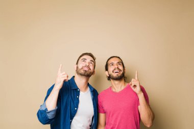 Happy handsome man hugging his gay boyfriend while pointing up to promote an ad against a studio background with copy space