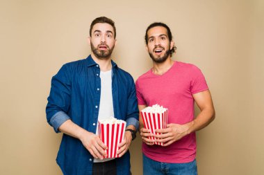 Excited young gay couple eating popcorn and looking surprised while watching a movie during a date