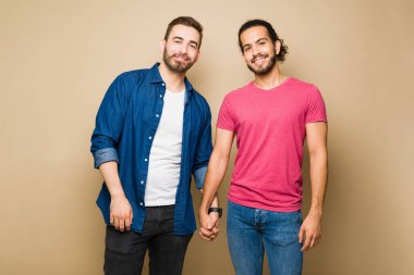 Attractive multiracial gay couple holding hands smiling while looking at the camera in front of a studio background