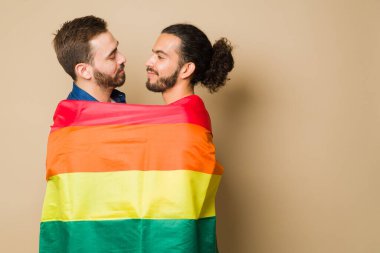 Romantic gay couple wrapped in a rainbow flag supporting the LGBT community rights while embracing against a background with copy space