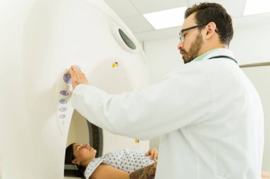 Caucasian male radiologist seen from behind starting the machine to do a CAT scan or tomography on a female patient