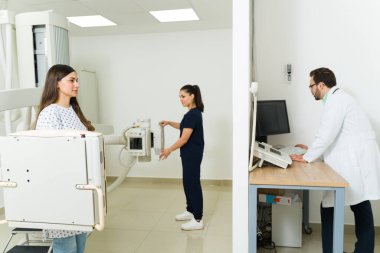 Hispanic young woman at the medical imaging lab getting an x-ray test of her back and chest with the help of a technician and radiologist 