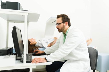 Side view of a caucasian male radiologist looking at the computer while doing a densitometry test on a young woman at the lab