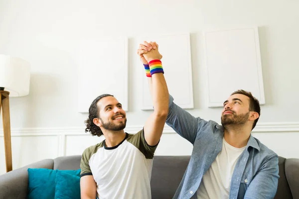 Happy queer men wearing rainbow wristbands holding hands as a couple and raising their hands to support LGBT gay rights