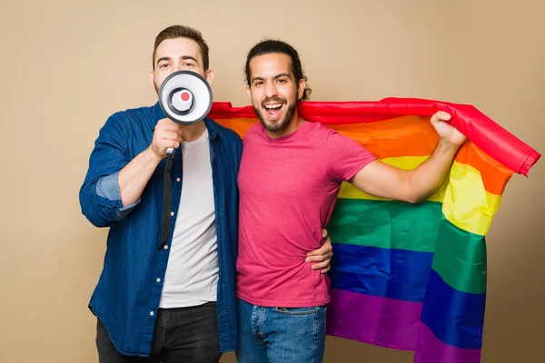 Excited caucasian man and latin boyfriend screaming using a megaphone during a LGBT protest to support gay rights with a rainbow flag