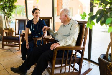Happy beautiful nurse at the nursing home laughing and chatting with a retired old man relaxing on a rocking chair