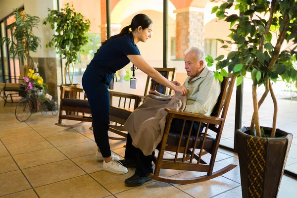 Caring female latin nurse working at the nursing home giving a warm blanket to an old man sitting on a rocking chair