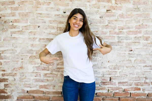 一位美丽的惊慌失措的女人站在室外的砖墙前 用自己设计的标志来展示自己的造型白色T恤 — 图库照片