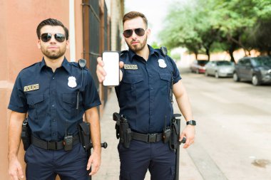 Üzgün erkek polis memurları GPS uygulama teknolojisiyle bir suçluyu takip ediyorlar ve akıllı telefon ekranını mesajla gösteriyorlar. 