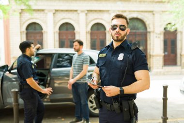 Siyah üniformalı ve güneş gözlüklü bir polis memuru trafik cezası yazarken kameraya bakıyor.