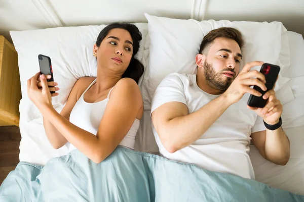 Jealous hispanic woman in bed spying on his happy partner or husband while texting on the phone and cheating