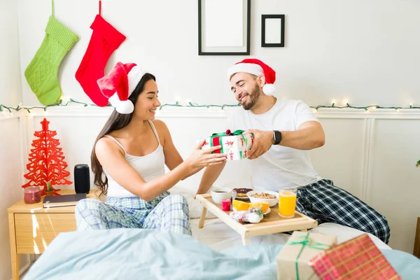Cheerful young man in pajamas giving a christmas present to his wife or girlfriend after waking up in the bedroom
