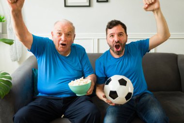 Heyecanlı yaşlı baba ve beyaz adam evde futbol maçı izlerken sevinçle bağırıyor ve tezahürat yapıyor.