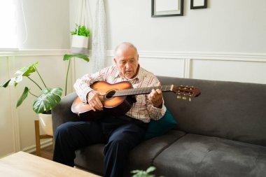 Yaşlı yetenekli bir adam kanepede otururken gitar çalıp şarkı söylüyor.