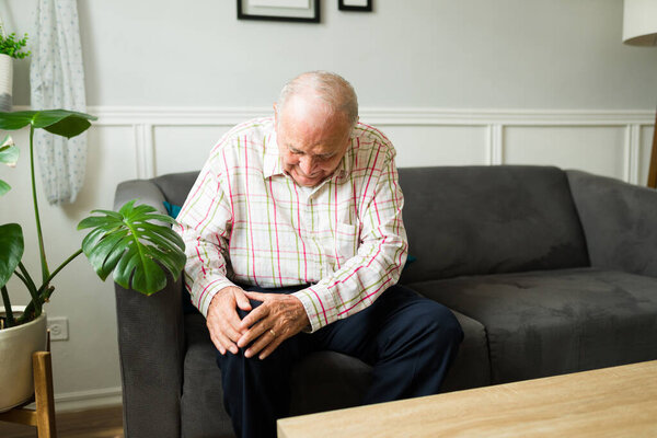 Upset old man looking sick suffering from knee or joint pain while resting on the sofa alone at home