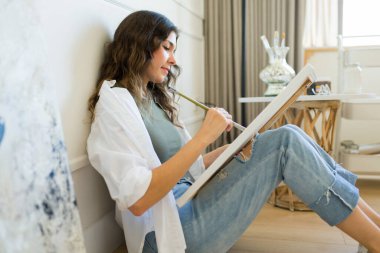 Rahatlamış genç bir kadın ressamın yan görüntüsü yerde rahatlıyor ve bir sanat eseri çizerken mutlu görünüyor.