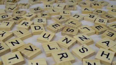 Harfler beyaz tahtaya rastgele dağılmış. Kamera geriye doğru gidiyor. Beyaz desenli bir yüzeye rastgele dağılmış ahşap karolar. Scrabble heceleme oyunu.