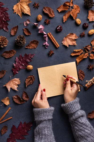Autumn fall thanksgiving day composition with decorative dried leaves. Female hands writing in notebook. Flat lay, view from above, still life seasonal background for greeting card, copy space