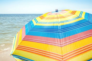 Mavi gökyüzü ve deniz zemininde parlak renkli şemsiye. Yaz tatilleri, dalgalar ve deniz yıkama kıyıları. Yaz manzarasında plaj gereksinimleri