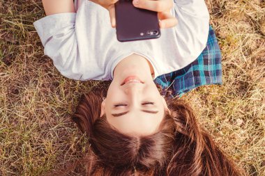 Bu fotoğraf parkta çimlerin üzerinde sırt üstü uzanmış ve cep telefonunu kullanan güzel bir genç kızın görüntüsünü tasvir ediyor. Uzak çalışma ve doğada serbest çalışma, üst görünüm.