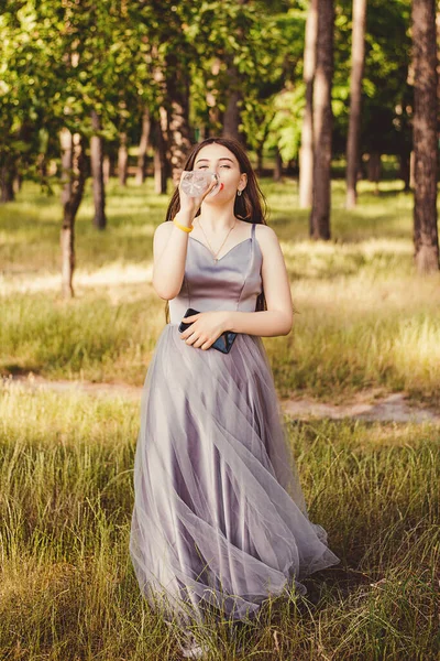 この写真は 夏の緑の公園で水を飲むと灰色のふわふわのドレスを着た若い美しい髪の少女の肖像画のイメージを示しています — ストック写真