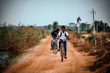 Davangere, Karnataka, Hindistan - 19 Şubat 2021: Güney Hint köyü çocukları (arkadaşları) yakındaki bir köyde çamurlu bir yolda bisikletle (bisikletlerle) okula gidiyorlar.