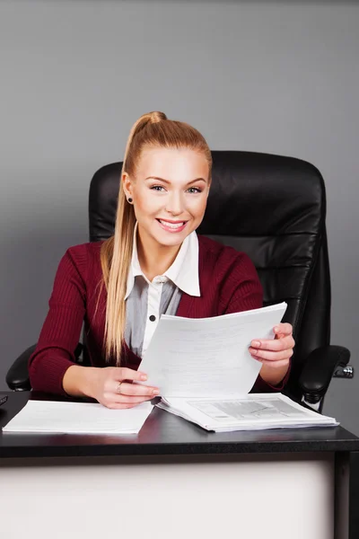 Mujer de negocios sonriente sentada en el lugar de trabajo y leyendo papel en la oficina —  Fotos de Stock