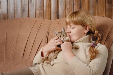 Woman in beige sweater sitting on sofa hugging cute tabby kitten indoors from low angle view with copy space