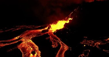 Drone capturing closely an earth's natural phenomenon. The shot shows erupting fountain of boiling red hot lava which flows in thick rivers over Geldingadalir valley in Iceland glowing in the dark night and dense clouds of ash ascending.