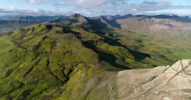 Groups of white geese cross paths on the water's surface. The geese move their wings and fly at a very low altitude as others stay on beach land. The beach is very large and has black volcanic rocky earth. white clouds reaching the horizon.
