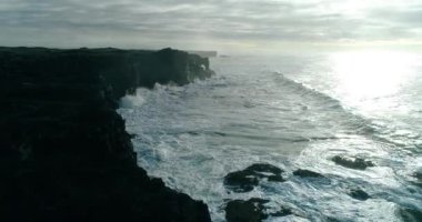 Güçlü rüzgarlar büyük deniz dalgalarını kayalık bir kıyıya doğru itiyor. Kıyı, volkanik siyah taştan oluşmuş muazzam bir yüksek arazidir. Atış, bulutlu bir kış gününde kıyı şeridiyle paralel uçan bir dron tarafından yapıldı.