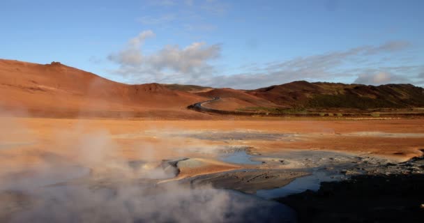 从冰岛的通风口和泥点排放出的硫磺烟在棕色的平原中央一大片灰色泥点不断地喷出硫磺烟 背景是两座覆盖着棕色土壤的山 其中一个有绿色的草皮 — 图库视频影像